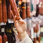 Woman chooses dried smoked sausage hanging in refrigerator in butcher’s shop